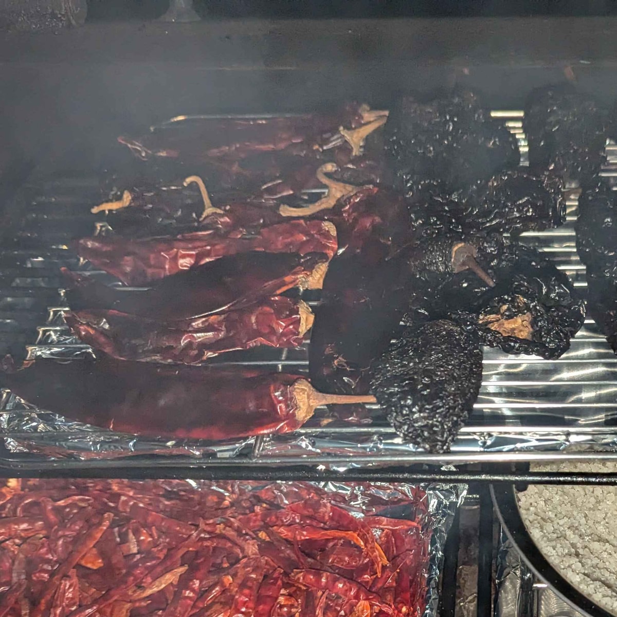 Red chiles and darkly roasted chiles lying together on the smoker rack, in smoke.